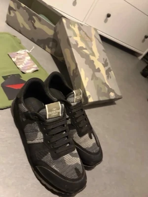 Valentino rockrunners camo sneakers i svart/grå - Snygga sneakers från Valentino med svart och grått camomönster. Skorna har svarta snören, meshpaneler och detaljer i mocka. Klassisk låg modell med robust sula och logga på plösen. Perfekta för dig som vill sticka ut med stilrena detaljer.