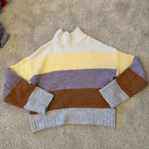 Randig stickad tröja Gina Tricot - Mjuk stickad tröja från Gina Tricot med breda ränder i vit, gul, lila, brun och ljusgrå. Tröjan har hög krage, avslappnad passform och ribbade muddar vid ärmslut och nederkant. Tröjan har gått ur sortimentet.