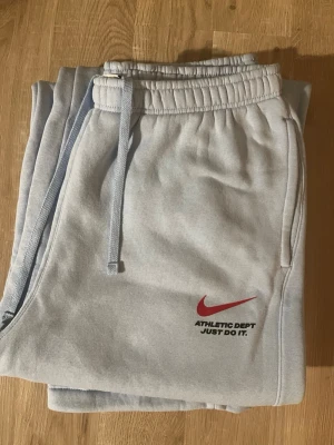 Ljusblå Nike mjukisbyxor - Snygga ljusgrå mjukisbyxor från Nike med röd logga och texten 'ATHLETIC DEPT JUST DO IT!' på vänster ben. Byxorna har resår och dragsko i midjan samt sidofickor. Perfekta för en avslappnad stil och tillverkade i mjukt material.
