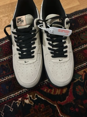 Nike Air Force 1 vita och svarta sneakers - Storlek 45 Nike Air Force 1 sneakers i vitt mockaliknande material med svarta detaljer och svarta skosnören. Klassisk låg modell med perforerad tå och Nike-logga på plös, häl och innersula. Snygg kontrast mellan den ljusa ovandelen och den svarta sulan.