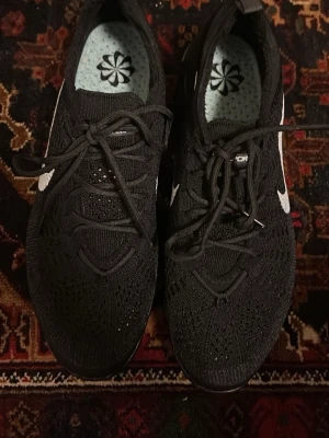 Svarta Nike Flyknit sneakers - Storlek 45. Svarta Nike Flyknit sneakers med vit swoosh på sidan. Skorna har stickad ovandel i mesh för extra andningsförmåga och svarta skosnören. Sulan är låg och innersulan har ett blommönster. Perfekta för en sportig och stilren look.