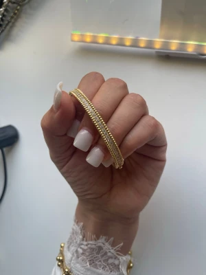 18k Guldpläterad armband med pärldetaljer - Snyggt armband i guldfärgad metall med små pärldetaljer längs hela bandet. Stilren och elegant design som passar perfekt till många olika looks. Armbandet är stelt och har en klassisk form som gör det lätt att matcha med andra smycken.