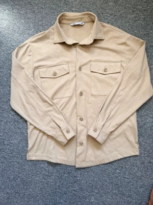 Beige overshirt från Zara - Säljer en beige overshirt från Zara med klassisk krage, två stora bröstfickor med knappar och långa ärmar. Materialet är mjukt och känns skönt mot huden. Hör av dig vid funderingar eller behovs av fler bilder!🤗