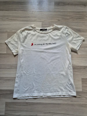 Vit t-shirt med text från Bik Bok - Vit t-shirt från Bik Bok med broderad text 'I'm looking for my other half.' och ett rött hjärta på bröstet. Klassisk passform med rund hals och korta ärmar. Tillverkad i mjuk bomull som känns skön mot huden. Den har en fläck i nedre kant fram och bak.  Har ej försökt få bort den.