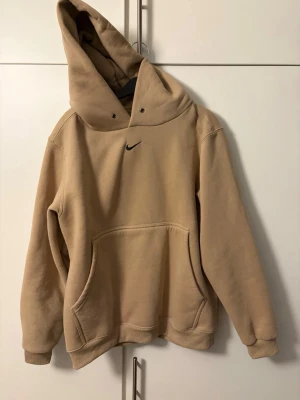 Beige hoodie från Nike med huva - Säljer en beige hoodie från Nike med klassisk svart swoosh-logga på bröstet. Hoodien har stor känguruficka framtill, ribbade muddar och en rejäl huva. Perfekt för en chill och avslappnad stil. Materialet känns mjukt och skönt mot huden.