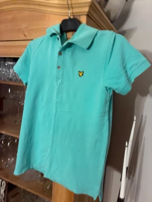 Turkos pikétröja från Lyle & Scott - Snygg turkos pikétröja från Lyle & Scott med klassisk krage och knappslå framtill. Tröjan har korta ärmar och den ikoniska gula örnloggan broderad på bröstet. Perfekt till jeans eller shorts för en clean och fräsch look. Använd ett fåtal gånger och har storleken M