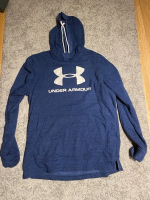 Blå hoodie från Under Armour - Snygg blå hoodie från Under Armour med stor vit logga och text på bröstet. Hoodien har huva med vita snören och en stor magficka framtill.