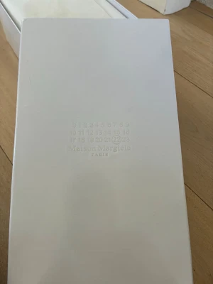 Vit skokartong från Maison Margiela - Vit skokartong från Maison Margiela Paris med det klassiska numrerade logotyptrycket på locket. Perfekt för dig som vill ha en snygg och stilren förvaring till dina skor eller sneakers.