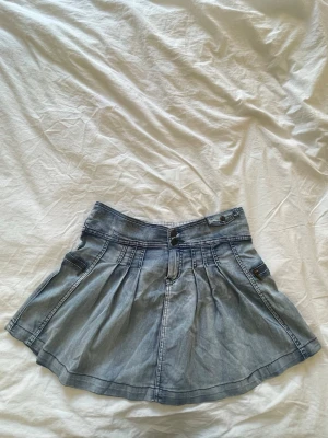 Ljusblå jeanskjol med fickor - Snygg ljusblå jeanskjol med utställd form och markerade sömmar. Kjolen har två fuskfickor fram och bak samt ett dekorativt bälte bak i midjan. Perfekt för dig som gillar en avslappnad och trendig stil.