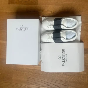 Valentino Garavani vita sneakers - Snygga vita sneakers från Valentino Garavani med bred svart rem över vristen och diskret logga. Skorna har klassisk låg siluett, rund tå och är tillverkade i läder. Kommer med originalkartong och dustbag.