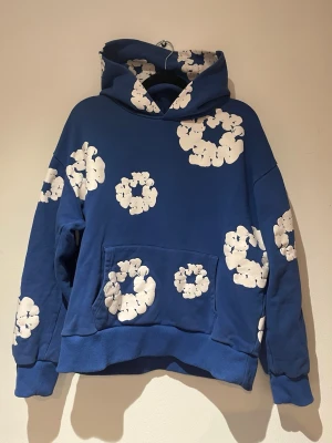 Denim tears - Cool Denim tear hoodie med stora vita blommönster över hela tröjan. Har en rymlig huva och en klassisk känguruficka framtill. Oversized passform och mjukt material som känns skönt mot huden. Perfekt för dig som gillar streetwear och unika prints.