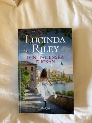 Den italienska flickan av Lucinda Riley - En gripande roman av Lucinda Riley med ett färgstarkt omslag som visar en kvinna i vit klänning vid en italiensk sjö. Boken är tryckt i mjukt omslag och handlar om kärlek, passion och hemligheter i Italien. Perfekt för dig som gillar romantik och dramatik.