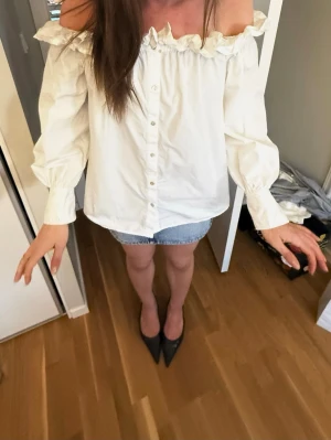 Vit off shoulder blus med volang - Trendig vit off shoulder blus med volangkant upptill och knappar framtill. Blusen har långa puffärmar med breda muddar och är gjord i ett lätt bomullsliknande material. Perfekt för en chic och avslappnad look.