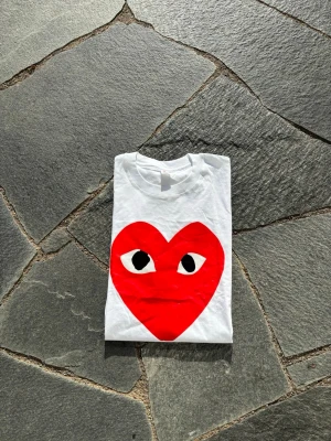COMME des GARÇONS - Vit t-shirt med stort rött hjärta och ögon på bröstet. Klassisk rund halsringning och korta ärmar. Perfekt för dig som vill sticka ut med en cool och lekfull design.