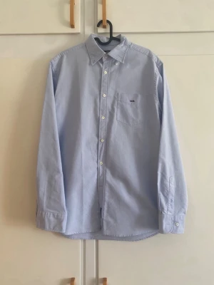 Ljusblå skjorta från POLO ASSN - Klassisk ljusblå skjorta från POLO ASSN med bröstficka och knappar framtill. Skjortan har lång ärm och stilren design, perfekt för en clean och snygg look. Tillverkad i bomull för en skön känsla.
