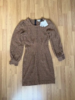 Rutigt brunt klänning från Second Female - Snygg brun- och svartmönstrad klänning från Second Female med rutigt mönster. Klänningen har långa puffärmar med breda manschetter och figurnära passform. Rund halsringning och dragkedja bak. Perfekt för dig som gillar retrostil.