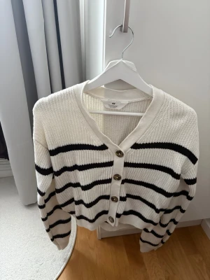 Vit kofta med svarta ränder från H&M - Snygg ribbstickad kofta från H&M i vitt med svarta horisontella ränder. Koftan har v-ringning, knappar framtill och långa ärmar. Perfekt för lager-på-lager och enkel att matcha med jeans eller kjol.