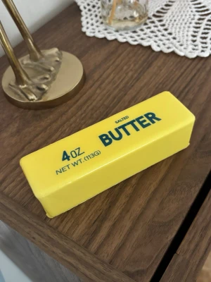 Butter Squishy Gul smörformad pryl i plast - Squishy leksak. Rolig pryl formad som ett smörpaket, gjord i gul plast med blå text. På sidan står det '4 OZ BUTTER SALTED'. Perfekt som dekoration eller skämtpresent till någon som gillar unika saker.