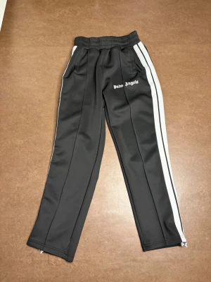 Palm Angels svarta mjukisbyxor - Palm Angels svarta mjukisbyxor med vita sidoränder och logga på vänster lår. Byxorna har resår i midjan, raka ben och dragkedja vid benslut. Tillverkade i ett glansigt, sportigt material som ger en trendig streetwear-look.