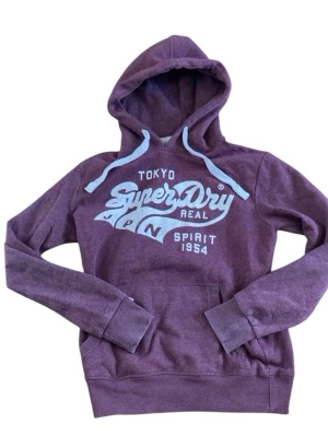 Vinröd  Superdry hoodie med tryck - Snygg vinröd hoodie från Superdry med stort vitt tryck på bröstet där det står 'Tokyo Superdry Real JPN Spirit 1954'. Hoodien har huva med vita dragsnören och en klassisk magficka. Perfekt för en avslappnad och trendig stil.