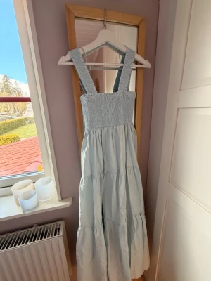 Ljusblå långklänning med breda axelband - Ljusblå långklänning med breda axelband och smockad överdel. Klänningen har en luftig och flowy passform med volanger i kjolen, perfekt för varma dagar. Tillverkad i ett mjukt och lätt material som känns skönt mot huden.