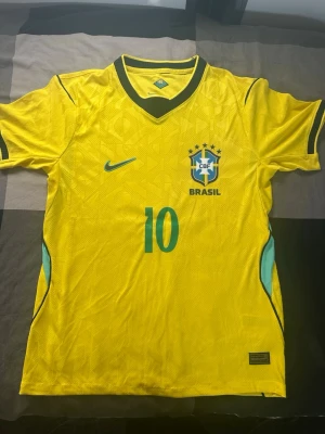 Gul Brasilien Nike fotbollströja - Snygg gul Brasilien fotbollströja från Nike med gröna och blå detaljer, nummer 10 på framsidan och CBF-emblemet broderat på bröstet. Tröjan har korta ärmar, V-ringning med grönt kantband och är tillverkad i ett lätt, ventilerande material perfekt för träning eller match.