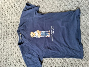 Blå Polo Ralph Lauren t-shirt med björn - Mörkblå t-shirt från Polo Ralph Lauren med klassisk björn-tryck på framsidan. T-shirten har rund halsringning och korta ärmar. Tillverkad i mjuk bomull, perfekt för en avslappnad och stilren look.