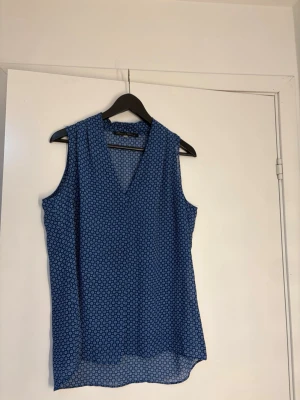 Blå ärmlös blus med mönster - Snygg blå ärmlös blus med v-ringning och små cirkelformade mönster i vitt. Luftigt material som passar perfekt till varma dagar. Lätt att matcha med både jeans och kjol för en avslappnad look.