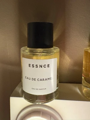 Essnce Eau de Caramel parfym - En parfymflaska från Essnce med doften Eau de Caramel. Flaskan är cylindrisk i transparent glas med en svart, matt rund kork. Innehållet är ljusgult och etiketten är vit med svart text. Perfekt för dig som gillar söta och karamelliga dofter.