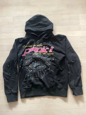 Sp5der hoodie PINK! - Svart sp5der hoodie med stor huva och färgglatt tryck på framsidan med spindelnät och texten 'Pink!'. Detaljer i rosa, gult och blått ger ett unikt uttryck. Perfekt för dig som gillar statement-plagg och streetstyle.