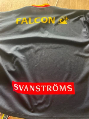 Djurgården IF hockeytröja Falcon - Djurgården IF hockeytröja i svart med gula och röda detaljer. Stort DIF-emblem på framsidan, sponsorer som Svanströms och EDS tryckta. Materialet är polyester och har en sportig passform, perfekt för hockeyfans.