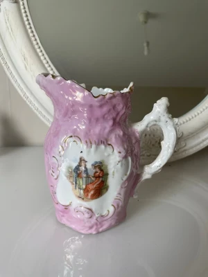 Unik dekorativ porslinskanna - Säljer en rosa dekorativ porslinskanna med vit och guld detaljering samt romantiskt motiv på framsidan. Kannans form är snirklig med vågig kant och ett vitt, ornamenterat handtag. Perfekt som prydnad eller samlarobjekt.