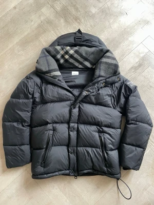 Svart Burberry jacka  - Säljer billigt för det blir varmare nu.Svart dunjacka från Burberry med klassiskt rutigt foder i huvan. Jackan har dragkedja och knappar framtill, två snedställda fickor med dragkedja och justerbar nederkant. Perfekt för kalla vinterdagar och riktigt snygg att bära.