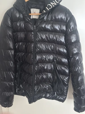 Svart dunjacka Moncler - Snygg svart dunjacka från Moncler med glansig finish och vadderad design. Jackan har dragkedja framtill, två fickor med dragkedja och en huva med Moncler-logga på insidan. Perfekt för kalla dagar och ger en trendig look. Innerfickan är sönder men går att sy ihop med några stygn