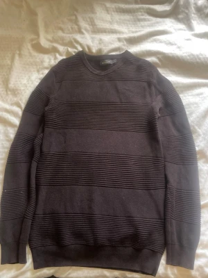 Brun tröja från Dressmann - Mörk brun  stickad tröja från Dressmann med breda horisontella ränder i olika stickstrukturer. Tröjan har rund halsringning och långa ärmar. Perfekt för lager-på-lager och enkel att matcha med jeans eller chinos.
