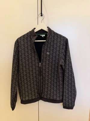 Mönstrad ziptröja från Lacoste - Säljer en grå ziptröja från Lacoste med grafiskt mönster i vitt och svart. Tröjan har ribbade muddar, ståkrage och dragkedja framtill. Klassisk Lacoste-logga på bröstet. Perfekt för en chill och stilren look.
