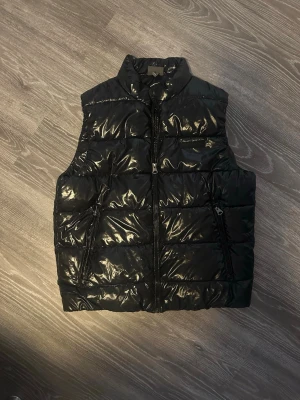 Svart glansig dunväst från H&M - Svart dunväst från H&M med glansig finish och hög krage. Västen har två snedställda fickor med dragkedja och quiltad design. Perfekt för lager-på-lager och ger en cool streetstyle-vibe.