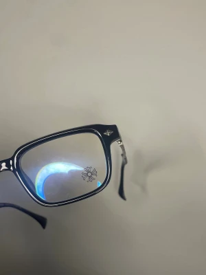 Chrome Heart glasögon  - Snygga svarta solglasögon med fyrkantiga bågar och mörka glas. Skalmarna har detaljer i silver som ger en cool kontrast. Perfekta för dig som vill ha en stilren och trendig look. Passar till alla outfits och ger en edgy vibe.
