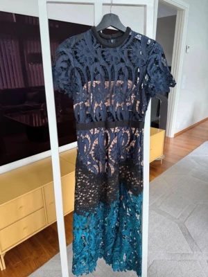 Blå och turkos spetsklänning från Self-Potrait - Unik långklänning i mörkblå och turkos spets med korta ärmar och rund hals. Klänningen har ett genombrutet mönster och markerad midja. Perfekt för dig som vill sticka ut med en elegant och detaljerad look.