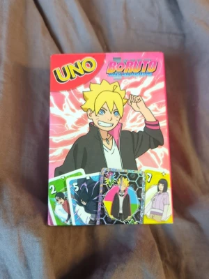 UNO Boruto Naruto Next Generations kortspel - UNO Boruto Naruto Next Generations kortspel med färgglada kort och animeinspirerade motiv från Boruto-serien. Kortleken har en rosa och gul design på förpackningen och innehåller flera karaktärer från serien. Perfekt för dig som gillar anime och samlarkortspel.