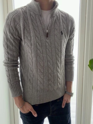 Grå kabelstickad tröja Polo Ralph Lauren - Snygg grå kabelstickad tröja från Polo Ralph Lauren med halv dragkedja och hög krage. Tröjan har ribbade muddar och nederkant samt den klassiska loggan broderad på bröstet. Perfekt för lager på lager och en clean look. Jag är 178 på bilden 