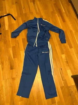 Blått tracksuit-set från Palm Angels - Snyggt blått tracksuit-set från Palm Angels med vita detaljer och dragkedjor. Setet består av en zip-jacka med fickor och matchande byxor med vita ränder längs sidorna. Palm Angels-logga broderad på både jacka och byxa. Perfekt för chill eller streetwear.