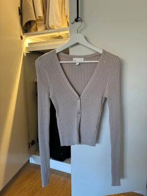 Beige ribbad kofta med v-ringning - Säljer en beige ribbad kofta med djup v-ringning och två knappar framtill. Koftan har lång ärm och är figurnära i modellen, perfekt att styla med jeans eller kjol. Materialet är stretchigt och mjukt för en bekväm känsla.