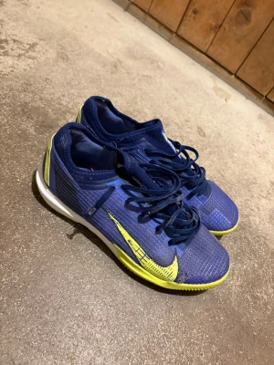 Nike Mercurial blå fotbollsskor - Säljer ett par Nike Mercurial fotbollsskor i blått med gula detaljer och vit sula. Skorna har snörning och är tillverkade i ett lätt syntetmaterial med texturerad yta för bättre bollkontroll. Perfekta för dig som vill ha snabbhet och stil på planen.