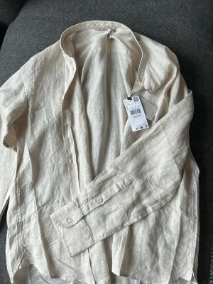 Beige linneskjorta från Mango - Stilren beige skjorta från Mango i 100% linne. Skjortan har klassisk krage, långa ärmar med knapp vid manschetten och en avslappnad passform. Perfekt för en fräsch och enkel look.