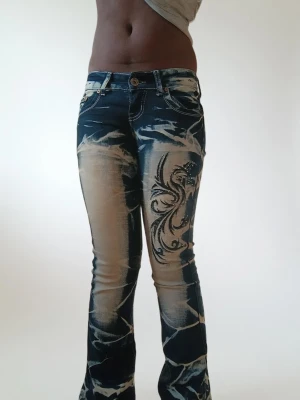 Crazy age bootcut jeans med mönster - Säljer ett par Crazy age jeans i bootcut-modell med coolt blekt mönster och broderade detaljer med svarta rosor och strass på både fram- och baksida. Jeansen är blå med ljusa partier och har låg midja. Perfekta för dig som gillar unik streetstyle. Har några fläckor som är sällsynta