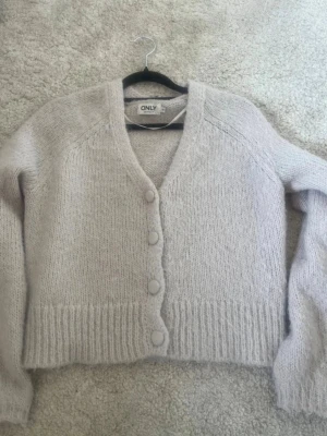 Ljusbeige kofta från ONLY - Mysig ljusbeige stickad kofta från ONLY med v-ringning och stora matchande knappar framtill. Koftan har lång ärm och ribbstickade muddar vid ärmslut och nederkant. 