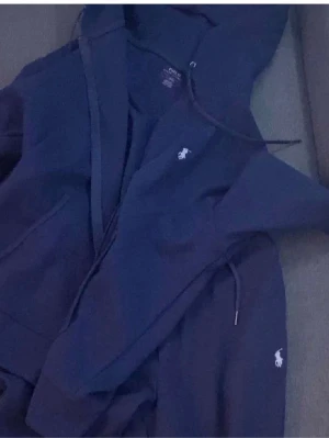 Mörkblå tracksiut från Polo Ralph Lauren - Mörkblå hoodie från Polo Ralph Lauren med klassisk huva och dragsko. Ikoniska vita polospelar-loggan broderad på bröstet. Mjuk bomullskänsla och känguruficka framtill. Perfekt för en avslappnad och stilren look.