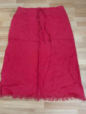 Röd/rosa långkjol med fransar och knyt - Säljer en röd/risa långkjol i linne med knytband i midjan och fransad nederkant. Kjolen har en enkel, rak passform och dragkedja bak. Perfekt för dig som gillar en avslappnad men ändå snygg stil. Midjan 88cm.