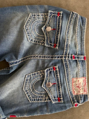 Blå True Religion jeans med kontrastsömmar - Säljer ett par blå jeans från True Religion med tydliga vita kontrastsömmar och röda detaljer. Jeansen har två bakfickor med lock och silverfärgade knappar samt en patch med logga bak i midjan. Klassisk bootcut-modell och slitningar för en cool look.🌷